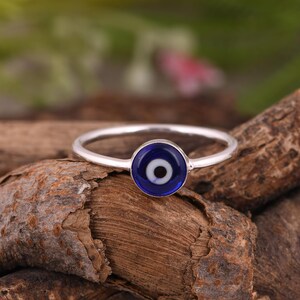 Puede incluir: Un anillo de plata con un dije circular que presenta un diseño de ojo en colores azul, blanco y negro. La banda es delgada y metálica, sosteniendo la pieza central redonda de aspecto vítreo en el medio. El diseño está centrado.