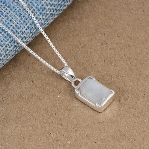 May include: A silver necklace featuring a rectangular pendant with a smooth, translucent white moonstone set in a polished silver frame. The pendant hangs from a delicate silver box chain, showcasing a clean, metallic finish and simple design.