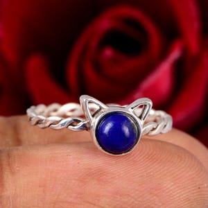 Puede incluir: Un anillo de plata con diseño de orejas de gato y una piedra redonda de color azul oscuro. La banda tiene un patrón de cuerda trenzada. El anillo se muestra sobre un fondo rojo borroso.