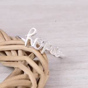 May include: A polished silver ring featuring the word hope written in a continuous cursive script. The metal band is thin and smooth, forming the letters of the word as the focal point of the jewelry piece, showcasing a reflective metallic finish.