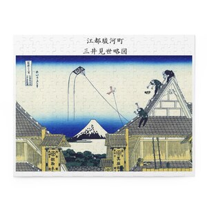 以下が含まれることがあります： 日本の木版画を特徴とするジグソーパズル。このアートワークは、屋根、人物、空を飛ぶ凧のある村のシーンを描いています。背景には山と青い空が描かれています。日本語のテキストが表示されます。