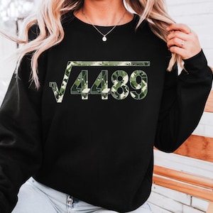 May include: Black sweatshirt with a square root symbol over the number 4489. The numbers are filled with a green and beige camouflage pattern. The sweatshirt has long sleeves and a crew neck.