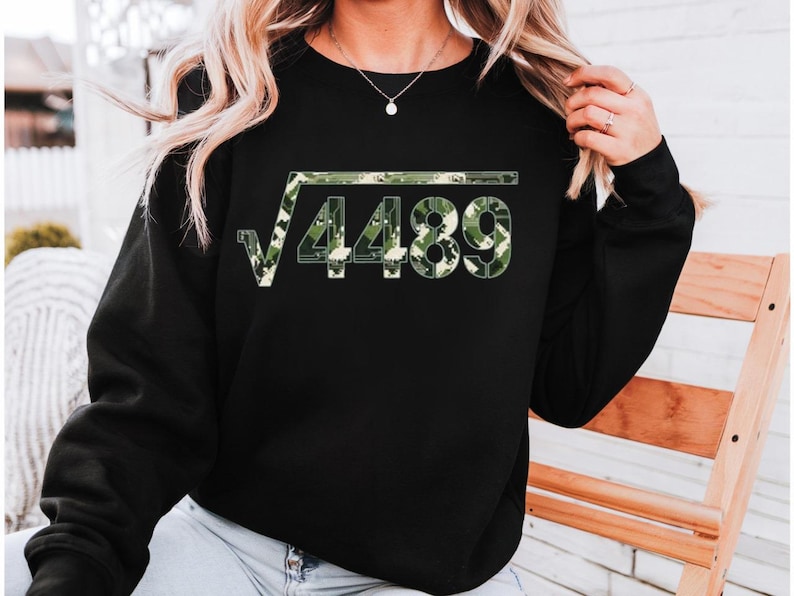 May include: Black sweatshirt with a square root symbol over the number 4489. The numbers are filled with a green and beige camouflage pattern. The sweatshirt has long sleeves and a crew neck.