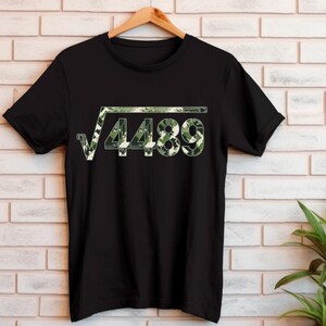 May include: Black t-shirt featuring a green camouflage design of a square root symbol and the number 4489. The shirt is displayed on a wooden hanger against a white brick wall.