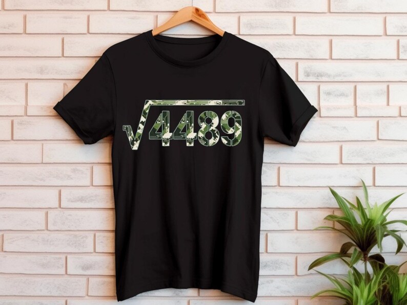 May include: Black t-shirt featuring a green camouflage design of a square root symbol and the number 4489. The shirt is displayed on a wooden hanger against a white brick wall.