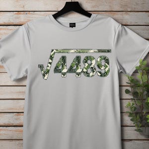 May include: A light grey t-shirt with a square root symbol over the number 4489. The numbers and symbols are in a camouflage pattern of green, brown, and white. The shirt is hanging on a black hanger.