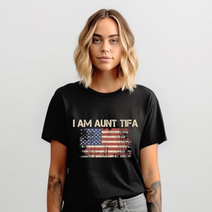 Könnte beinhalten: Schwarzes T-Shirt mit dem Text "I AM AUNT TIFA" über einem abgenutzten amerikanischen Flaggen-Design. Das T-Shirt wird von einer Person mit blonden Haaren und Tätowierungen am Arm getragen.