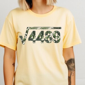 May include: A pale yellow t-shirt with a square root symbol and the number 4489 in a green and white camouflage design. The shirt has short sleeves and a round neck.