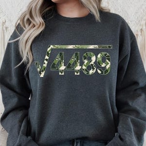 May include: Dark grey sweatshirt with a square root symbol and the number 4489 in a camouflage design. The pattern uses green, white, and tan. The sweatshirt is a casual garment.