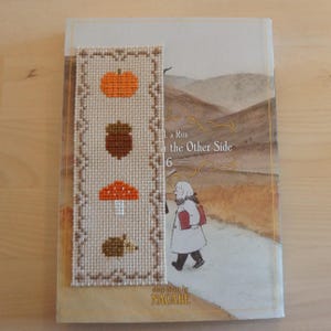 Puede incluir: Un marcapáginas hecho a mano con diseños de otoño: una calabaza, una bellota, una seta y un erizo. El marcapáginas está colocado sobre un libro titulado "a Run on the Other Side 6", con una ilustración de un niño caminando por un camino.