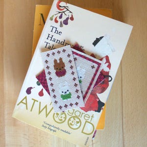 May include: A stack of books with a bookmark on top. The bookmark is cross-stitched with a brown bunny design and green stars. The book has the title "The Handmaid's Tale" and the author's name "Margaret Atwood".