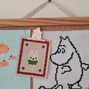 May include: A corkboard with cross-stitch art and a small print. The cross-stitch pieces feature a white cartoon character and a framed design with a bunny. A decorative clip with a cartoon character holds the framed cross-stitch.