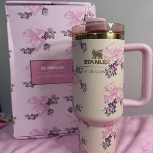 May include: A pink and cream-colored Stanley tumbler with a handle, adorned with pink bows and floral patterns. The tumbler has a gold rim and a pink lid. A matching box with the Stanley and Loveshackfancy logos is in the background.