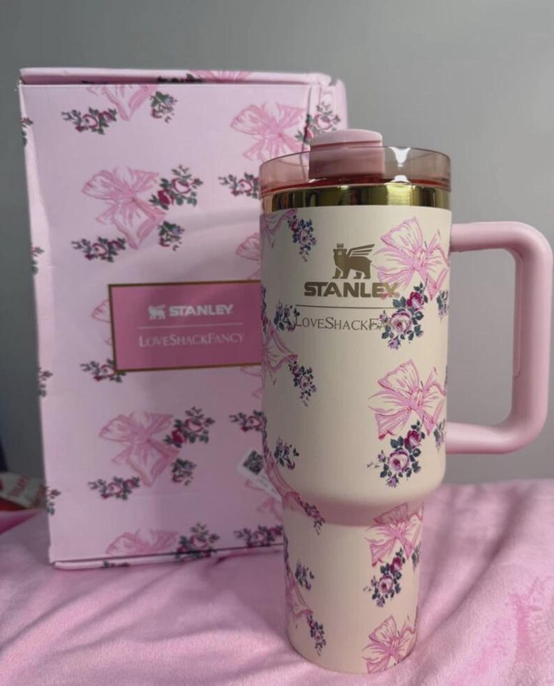 May include: A pink and cream-colored Stanley tumbler with a handle, adorned with pink bows and floral patterns. The tumbler has a gold rim and a pink lid. A matching box with the Stanley and Loveshackfancy logos is in the background.