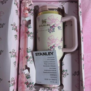 May include: A Stanley insulated mug with a handle, featuring a cream-colored body adorned with pink bows and floral patterns. The mug is packaged in a matching floral box with a pink straw. The Stanley logo is visible.
