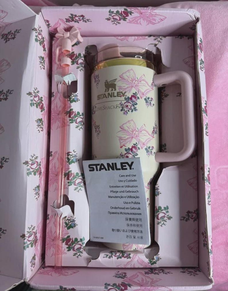 May include: A Stanley insulated mug with a handle, featuring a cream-colored body adorned with pink bows and floral patterns. The mug is packaged in a matching floral box with a pink straw. The Stanley logo is visible.