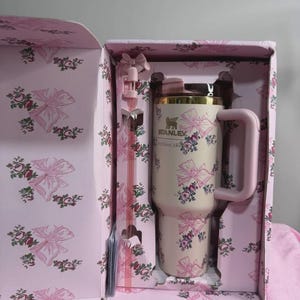 May include: A cream-colored Stanley tumbler with a pink floral and bow pattern, presented in a matching gift box. The tumbler has a pink handle and a gold rim. A pink straw is also included.