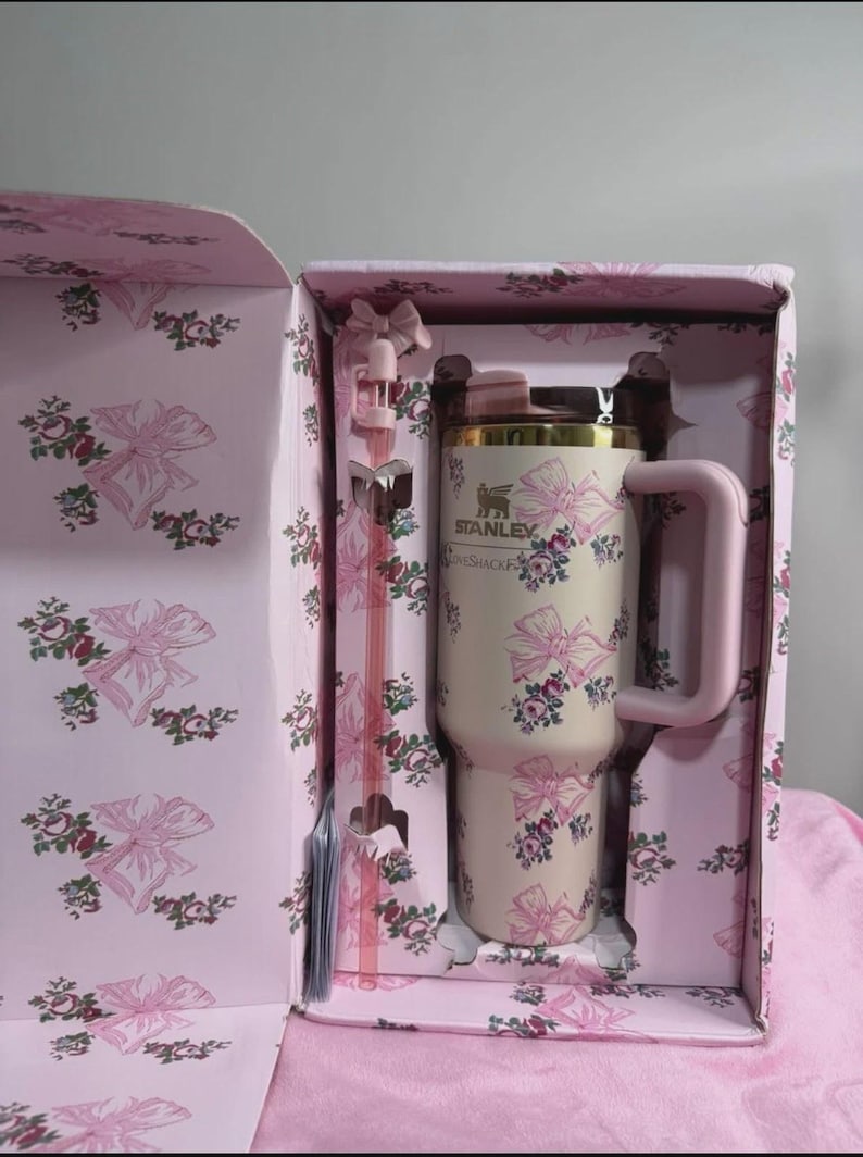 May include: A cream-colored Stanley tumbler with a pink floral and bow pattern, presented in a matching gift box. The tumbler has a pink handle and a gold rim. A pink straw is also included.