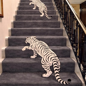 Puede incluir: Dos alfombras con forma de tigre blanco y rayas negras están colocadas sobre una escalera con moqueta gris oscuro. Las alfombras tienen forma de tigres en posición de caminar, con una alfombra más pequeña en un escalón alto y otra más grande.