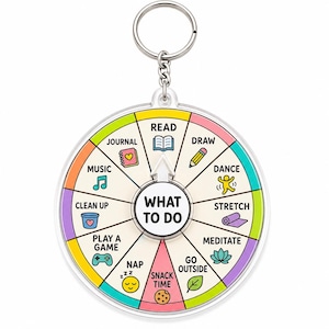 May include: A circular acrylic keychain featuring a spinning wheel labeled WHAT TO DO in the center. The wheel is divided into colorful segments with icons and text for activities including read, draw, dance, stretch, meditate, go outside, snack time, nap, play a game, clean up, music, and journal.