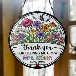 Può includere: Decorazione rotonda per finestre in vetro colorato con un design floreale e le parole "grazie" e "per avermi aiutato a crescere". L'opera d'arte include una varietà di fiori colorati, libellule e una farfalla. È incluso anche il nome "Mrs. Wilson".