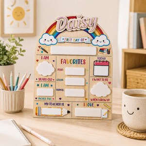Könnte beinhalten: Ein bogenförmiges Holzschild für den ersten Schultag mit dem Namen Daisy, einem Regenbogen und Wolken. Es enthält weiße Leerfelder zum Ausfüllen von Details wie Alter, Größe, Lieblingsessen, Farbe, Tier, Lehrer und zukünftigen Berufswünschen.