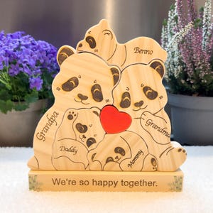 Könnte beinhalten: Holzskulptur einer Pandafamilie mit rotem Herz, mit den Worten "Grandpa", "Daddy", "Mommy" und "Grandma". Auf dem Sockel steht "We're so happy together". Die Pandas sind hellbraun mit schwarzen Akzenten.