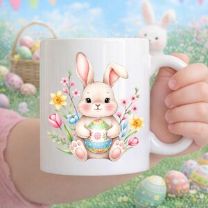 Puede incluir: Taza de cerámica blanca con un diseño de conejito de Pascua sosteniendo un huevo decorado, rodeado de flores coloridas. El diseño incluye flores rosas, amarillas y azules. La taza tiene un asa blanca y es sostenida por una persona.