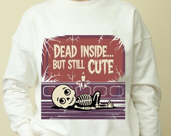 Peut inclure: Sweat-shirt blanc avec un dessin de squelette de dessin animé et le texte "DEAD INSIDE... BUT STILL CUTE". Le squelette est allongé avec une bougie et un fond décoratif. Le design utilise des couleurs rouge, violet et noir.