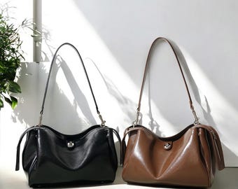 May include: Two leather shoulder bags, one black and one brown. Both have a curved design, long straps, and silver-tone metal hardware. They are set against a white backdrop, illuminated by natural light.