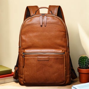 Pode incluir: Uma mochila de couro castanho com uma parte superior arredondada e um bolso frontal com zíper. A mochila tem duas alças de ombro ajustáveis e uma alça superior. O couro tem uma aparência texturizada. Um pequeno cacto num vaso de terracota é visível no fundo.