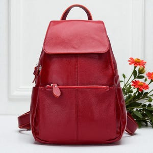 Puede incluir: Una mochila de cuero roja con cierre de solapa y un bolsillo frontal con cremallera. La mochila tiene correas ajustables y un asa superior. La bolsa está hecha de cuero texturizado y tiene herrajes plateados.