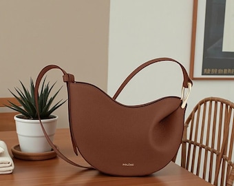 May include: A brown leather handbag with a curved design and a gold-coloured handle. The bag has a long strap and the word "POLÈNE" is printed on the front. The bag is sitting on a wooden table.