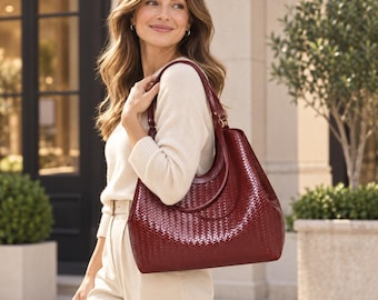 May include: A deep burgundy hobo bag featuring a woven leather texture and a curved top handle. The bag has a structured silhouette with a glossy finish and gold-toned hardware accents at the strap attachments, carried over the shoulder.