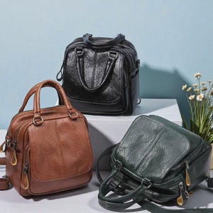 May include: Three leather handbags in black, brown, and green. Each bag features a top handle, a zipped main compartment, and a front pocket. The bags are displayed on a white surface against a light blue backdrop.
