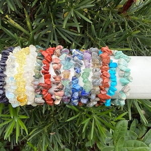 May include: A collection of colorful gemstone chip bracelets displayed on a white cylindrical object. The bracelets feature a variety of colors, including blue, yellow, red, green, and purple. The background is a lush green with foliage.