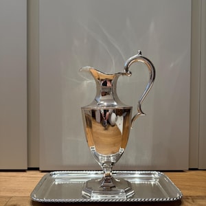 Może przedstawiać: Polerowany srebrny dzbanek na wodę z zakrzywionym uchwytem i wylewką, spoczywający na prostokątnej tacy z koralikową krawędzią. Dzbanek ma szeroką podstawę i rozkloszowaną górę. Taca i dzbanek odbijają otoczenie.