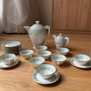 Può includere: Servizio da tè vintage con design verde chiaro e oro. Il set include una teiera, una lattiera, una zuccheriera, tazze, piattini e piccole ciotole. Il set è disposto su una superficie di legno.