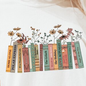 May include: Cream-colored t-shirt featuring a graphic of colorful books with floral accents. The book titles include "HOME ALONE", "POLAR EXPRESS", "LOVE ACTUALLY", and "The Grinch". The design is a whimsical representation of holiday classics.