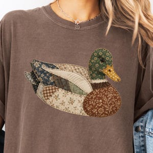 May include: Brown t-shirt featuring a patchwork duck design. The duck has a green head, yellow beak, and a body composed of various fabric patterns in shades of blue, brown, and cream. The shirt has rolled-up sleeves.