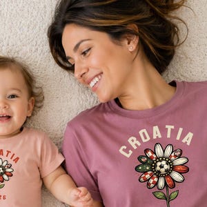 May include: A light pink t-shirt and a mauve t-shirt, both featuring a graphic of a flower with a soccer ball center, the text "CROATIA" arched above, and "2025" below. The flower petals incorporate red and white checkered patterns.