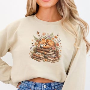 May include: A beige crewneck sweatshirt featuring a graphic print of a sleeping orange fox curled up on a stack of vintage books, surrounded by colorful wildflowers, ferns, butterflies, and a small cup of tea with a pair of glasses.
