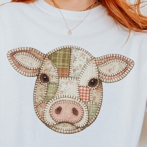 May include: A white short-sleeved t-shirt featuring a central graphic of a cow head designed with a patchwork pattern. The cow illustration incorporates various muted green, beige, and red floral and checkered fabric textures with dark stitching.