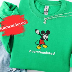 May include: A green embroidered sweatshirt featuring a cartoon Mickey Mouse character in a dabbing pose. The text "overstimulated" is below the character. A silver necklace is draped on the sweatshirt. Blue jeans are visible in the background.