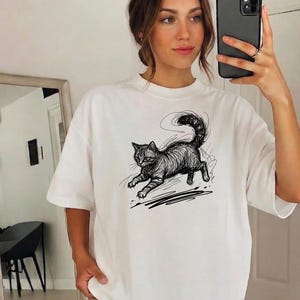 May include: A white short-sleeved t-shirt featuring a black ink-style sketch of a cat in motion. The cat is depicted with scribbled, expressive lines, showing a curved tail and a pouncing posture. The design is centered on the chest of the shirt.