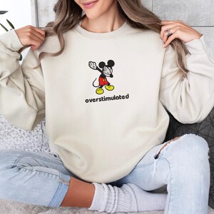 May include: Cream-colored sweatshirt featuring a cartoon Mickey Mouse graphic in a dabbing pose, with red shorts and yellow shoes. The word "overstimulated" is printed below the character in black.