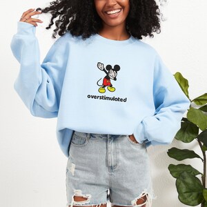 May include: Light blue sweatshirt featuring a cartoon Mickey Mouse character in a dabbing pose. The text "overstimulated" is printed below the character in a black font. The sweatshirt is a solid light blue color.