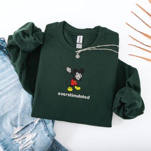 May include: A dark green sweatshirt with a Mickey Mouse graphic and the word "overstimulated" in white. The sweatshirt is paired with a silver necklace and blue jeans.