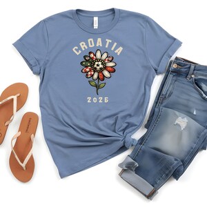 May include: A dusty blue short-sleeve t-shirt featuring a graphic of a flower with red and white checkered petals and a soccer ball center. The text CROATIA is arched above the flower, and the number 2026 is printed below it in a light cream color.