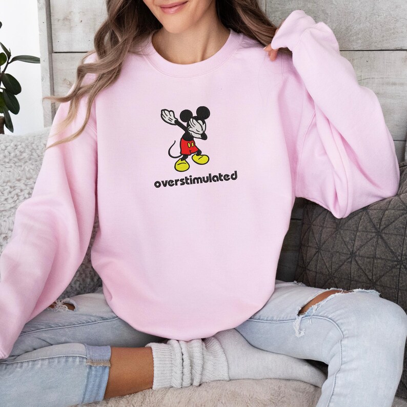 May include: A light pink sweatshirt featuring a cartoon Mickey Mouse character in a dabbing pose. The character is wearing red shorts, yellow shoes, and black gloves. The word "overstimulated" is printed in black below the character.